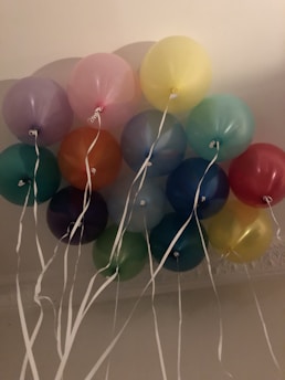 A cluster of colorful balloons with white strings attached, floating indoors against a light-colored ceiling.
