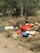 A pile of assorted garbage and debris, including colorful bags and a discarded stuffed toy, is scattered along a roadside surrounded by dry grass and trees.