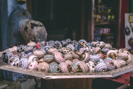 A collection of colorful ceramic drawer knobs is displayed on a large, ornate metal tray. The knobs exhibit a variety of patterns, including stripes, floral designs, and geometric shapes, and are arranged in a somewhat haphazard manner. The background features shelves filled with similar decorative items, adding to the artisanal marketplace ambiance.