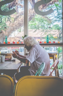 An elderly person with white hair is sitting at a wooden table in a cafe, engaged with something in their hands. The backdrop features large windows showcasing a view of trees and structures outside. Small vases or bottles and decorative items are arranged on the window ledge.