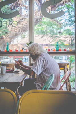 An elderly person with white hair is sitting at a wooden table in a cafe, engaged with something in their hands. The backdrop features large windows showcasing a view of trees and structures outside. Small vases or bottles and decorative items are arranged on the window ledge.