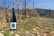 A bottle of wine with a blue cap and label stands on rocky soil between rows of dormant grapevines. The vineyard extends into the background, with hills visible under a clear blue sky.
