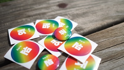 A collection of stickers and buttons featuring a rainbow gradient background with white text in a non-Latin script placed on a wooden surface.