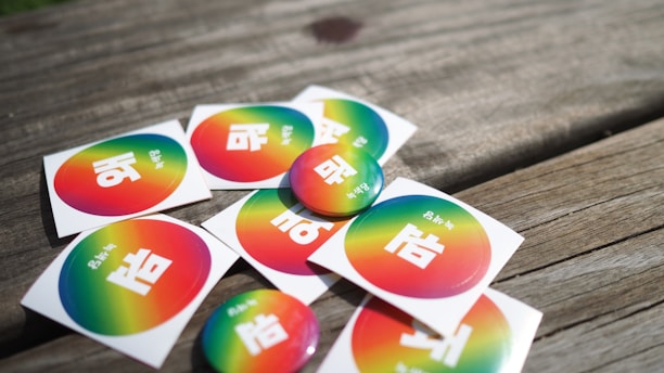 A collection of stickers and buttons featuring a rainbow gradient background with white text in a non-Latin script placed on a wooden surface.