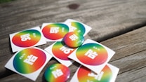 A collection of stickers and buttons featuring a rainbow gradient background with white text in a non-Latin script placed on a wooden surface.