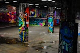 Vivid graffiti art covers multiple concrete pillars in an underground space, featuring a variety of colorful and intricate designs, including faces, abstract shapes, and lettering. The floor is reflective with small puddles, adding to the dynamic and urban atmosphere of the setting.