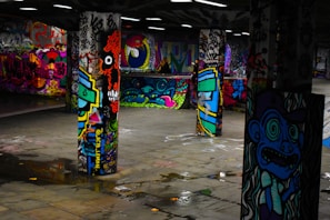 Vivid graffiti art covers multiple concrete pillars in an underground space, featuring a variety of colorful and intricate designs, including faces, abstract shapes, and lettering. The floor is reflective with small puddles, adding to the dynamic and urban atmosphere of the setting.