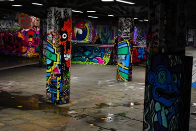Vivid graffiti art covers multiple concrete pillars in an underground space, featuring a variety of colorful and intricate designs, including faces, abstract shapes, and lettering. The floor is reflective with small puddles, adding to the dynamic and urban atmosphere of the setting.
