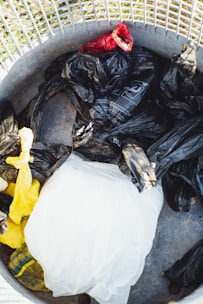 A collection of plastic bags in various colors, including black, yellow, and red, is placed inside a metal trash can. The surface of the can is visible, along with a metal grid at the top that likely forms part of its lid or a casing.