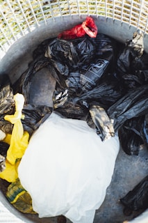 A collection of plastic bags in various colors, including black, yellow, and red, is placed inside a metal trash can. The surface of the can is visible, along with a metal grid at the top that likely forms part of its lid or a casing.