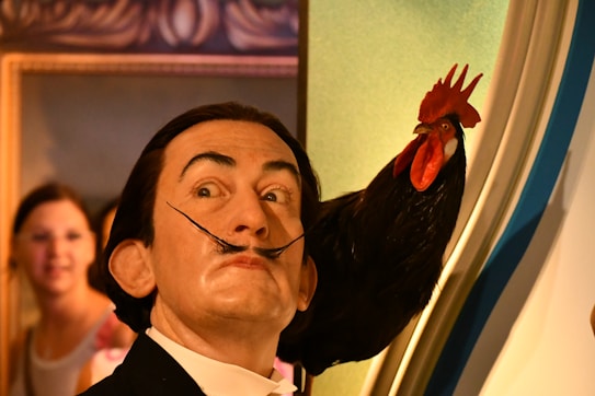 A wax figure of a man with an exaggerated mustache looks upward with a surprised expression. Beside him is a lifelike rooster with vibrant red comb and wattles. In the background, two people are visible, slightly out of focus, observing the scene. The setting appears to be an indoor exhibit with framed art visible behind.