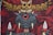 An artistic mural featuring a whimsical creature with large eyes and tusks holding two spears. Above this creature, an intricately detailed owl stretches its wings. The background is filled with red foliage-like patterns, adding a mysterious and fantastical element.