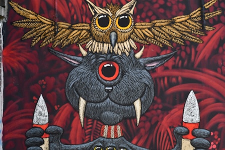 An artistic mural featuring a whimsical creature with large eyes and tusks holding two spears. Above this creature, an intricately detailed owl stretches its wings. The background is filled with red foliage-like patterns, adding a mysterious and fantastical element.