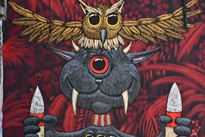 An artistic mural featuring a whimsical creature with large eyes and tusks holding two spears. Above this creature, an intricately detailed owl stretches its wings. The background is filled with red foliage-like patterns, adding a mysterious and fantastical element.