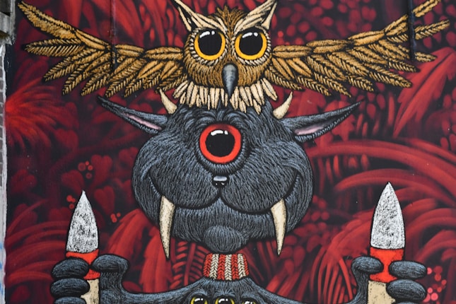 An artistic mural featuring a whimsical creature with large eyes and tusks holding two spears. Above this creature, an intricately detailed owl stretches its wings. The background is filled with red foliage-like patterns, adding a mysterious and fantastical element.