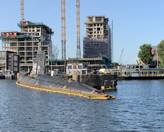 A partly submerged submarine is moored in a harbor area surrounded by water. Several construction cranes and partially built structures are visible in the background, indicating ongoing construction or development. The sky is clear, and some trees are present near the water and buildings.