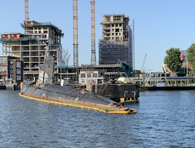 A partly submerged submarine is moored in a harbor area surrounded by water. Several construction cranes and partially built structures are visible in the background, indicating ongoing construction or development. The sky is clear, and some trees are present near the water and buildings.