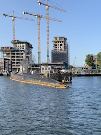 A partly submerged submarine is moored in a harbor area surrounded by water. Several construction cranes and partially built structures are visible in the background, indicating ongoing construction or development. The sky is clear, and some trees are present near the water and buildings.