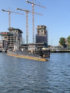 A partly submerged submarine is moored in a harbor area surrounded by water. Several construction cranes and partially built structures are visible in the background, indicating ongoing construction or development. The sky is clear, and some trees are present near the water and buildings.