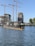 A partly submerged submarine is moored in a harbor area surrounded by water. Several construction cranes and partially built structures are visible in the background, indicating ongoing construction or development. The sky is clear, and some trees are present near the water and buildings.