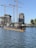 A partly submerged submarine is moored in a harbor area surrounded by water. Several construction cranes and partially built structures are visible in the background, indicating ongoing construction or development. The sky is clear, and some trees are present near the water and buildings.