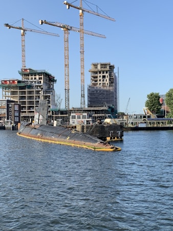 A partly submerged submarine is moored in a harbor area surrounded by water. Several construction cranes and partially built structures are visible in the background, indicating ongoing construction or development. The sky is clear, and some trees are present near the water and buildings.