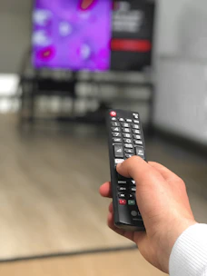 A hand holding a remote control in front of a TV showing sports and kids channels.