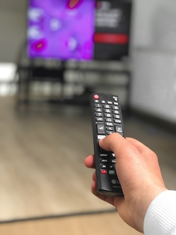 A hand holding a black remote control in a room with a blurred television screen in the background displaying purple and black colors.
