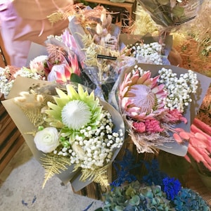 A collection of elegantly arranged flower bouquets, featuring a variety of blooms including proteas, baby's breath, and roses. The bouquets are wrapped in brown paper and displayed on a market stall with a mix of natural and artificial lighting, highlighting the intricate textures and colors of the flowers.