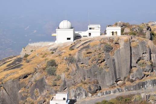 An observatory complex perched on a rocky hilltop with several white buildings, including a domed structure. Surrounding the buildings are rugged rocks and sparse vegetation with patches of dried grass.