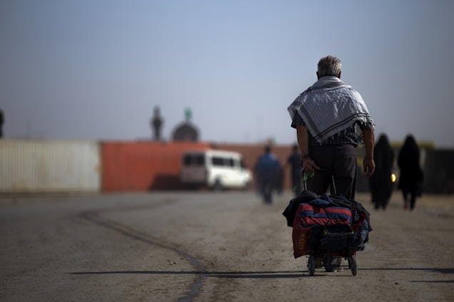 A man walks along a dusty road pulling a cart filled with bags. His back is turned to the camera, wearing a patterned shawl over his shoulders. Several other people in the background walk in the same direction. Off to the side, a vehicle is parked against a backdrop of shipping containers.