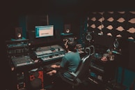 A person is seated in a professional recording studio, surrounded by various electronic equipment including a computer, multiple speakers, and audio mixers. The room is dimly lit, creating a focused and immersive atmosphere. The walls are lined with acoustic panels to optimize sound quality.