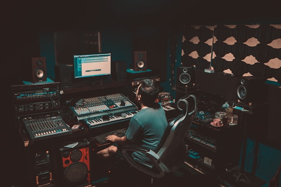 A person is seated in a professional recording studio, surrounded by various electronic equipment including a computer, multiple speakers, and audio mixers. The room is dimly lit, creating a focused and immersive atmosphere. The walls are lined with acoustic panels to optimize sound quality.