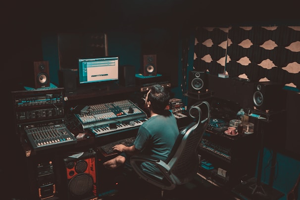 A person is seated in a professional recording studio, surrounded by various electronic equipment including a computer, multiple speakers, and audio mixers. The room is dimly lit, creating a focused and immersive atmosphere. The walls are lined with acoustic panels to optimize sound quality.