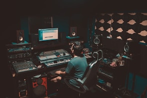 A person is seated in a professional recording studio, surrounded by various electronic equipment including a computer, multiple speakers, and audio mixers. The room is dimly lit, creating a focused and immersive atmosphere. The walls are lined with acoustic panels to optimize sound quality.
