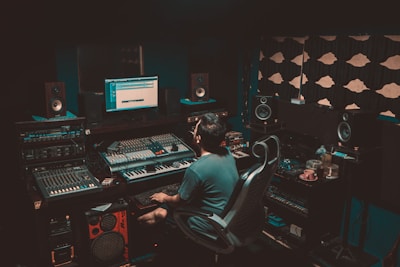 A person is seated in a professional recording studio, surrounded by various electronic equipment including a computer, multiple speakers, and audio mixers. The room is dimly lit, creating a focused and immersive atmosphere. The walls are lined with acoustic panels to optimize sound quality.