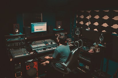 A person is seated in a professional recording studio, surrounded by various electronic equipment including a computer, multiple speakers, and audio mixers. The room is dimly lit, creating a focused and immersive atmosphere. The walls are lined with acoustic panels to optimize sound quality.