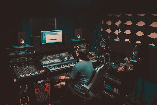 A person is seated in a professional recording studio, surrounded by various electronic equipment including a computer, multiple speakers, and audio mixers. The room is dimly lit, creating a focused and immersive atmosphere. The walls are lined with acoustic panels to optimize sound quality.