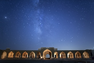 A starry night sky with the Milky Way galaxy visible above an ancient stone structure. The structure features multiple arched doorways illuminated by warm lights, creating a contrast with the deep blue of the night sky filled with stars.