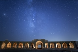 A starry night sky with the Milky Way galaxy visible above an ancient stone structure. The structure features multiple arched doorways illuminated by warm lights, creating a contrast with the deep blue of the night sky filled with stars.
