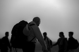 Several silhouettes of people are visible against a bright backdrop. One person, in the foreground, has a large backpack and looks to be heading in a direction opposite to the group in the background.