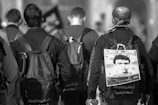 Three people are walking away from the camera. They are wearing dark clothing and carrying backpacks. One person has a poster attached to their backpack featuring a portrait of a man and some text. The scene appears to be outdoors in an urban setting.