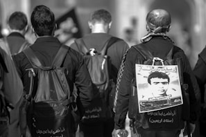 Three people are walking away from the camera. They are wearing dark clothing and carrying backpacks. One person has a poster attached to their backpack featuring a portrait of a man and some text. The scene appears to be outdoors in an urban setting.