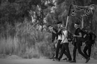 A group of people walking along a road, each carrying a backpack. One person is holding a flag with writing on it. The scene is in grayscale, indicating it might symbolize a journey or pilgrimage. They appear to be in a rural area with trees and vegetation in the background.