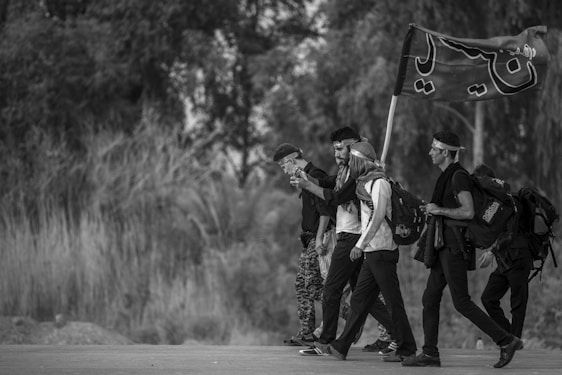 A group of people walking along a road, each carrying a backpack. One person is holding a flag with writing on it. The scene is in grayscale, indicating it might symbolize a journey or pilgrimage. They appear to be in a rural area with trees and vegetation in the background.
