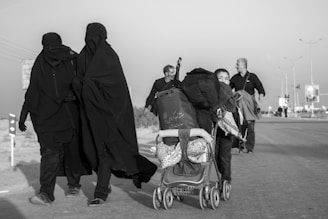 A black and white photo depicting a group of people on a road. Two women in black clothing walk closely together, while a man and a child are pushing a stroller filled with bags. The scene captures a sense of movement, possibly migration or pilgrimage, with additional people visible in the background.