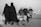 A black and white photo depicting a group of people on a road. Two women in black clothing walk closely together, while a man and a child are pushing a stroller filled with bags. The scene captures a sense of movement, possibly migration or pilgrimage, with additional people visible in the background.