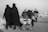 A black and white photo depicting a group of people on a road. Two women in black clothing walk closely together, while a man and a child are pushing a stroller filled with bags. The scene captures a sense of movement, possibly migration or pilgrimage, with additional people visible in the background.