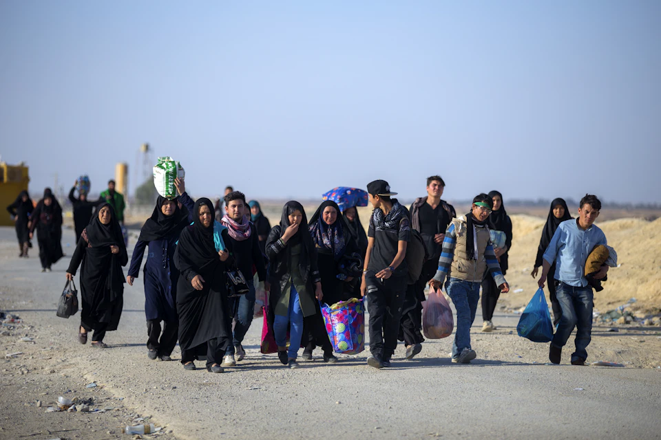 The image depicts a group of people walking down a dirt road, with some carrying bags and others holding umbrellas. The sky above them is clear blue, providing a beautiful backdrop to their journey. The perspective of the photo suggests it was taken from behind the group, giving an impression of being part of the crowd as they move forward together.

There are at least 12 people visible in the image, with some walking closer to the camera and others further away. They appear to be a diverse group, each carrying their own bags or umbrellas, indicating that they might be on a journey or traveling for various reasons. The overall atmosphere of the scene is one of camaraderie and shared purpose as these individuals move together down the road.