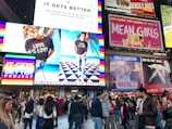 A bustling urban scene with large advertising billboards. The largest billboard is from American Eagle Outfitters, featuring models wearing t-shirts with 'Love is Love' text in rainbow colors, supporting the It Gets Better Project. Surrounding the main advertisement are other colorful billboards for shows, including 'Mean Girls' and 'Waitress'. A diverse crowd walks and takes photos below, adding to the vibrant city atmosphere.
